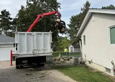 Tree removal crew lifting cut logs into truck with crane, suburban driveway
