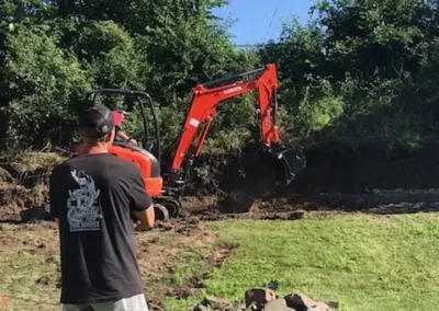 Worker operating orange mini excavator digging lawn near trees